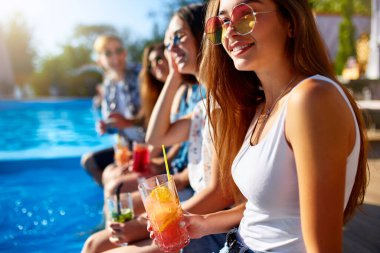 Güzel bir kadın, güneşli yaz gününde arkadaşlarıyla havuz kenarında taze renkli kokteyl bardağıyla rahatlar. İnsanlar tropikal tatillerde lüks havuz başı partisinde içki içiyorlar..