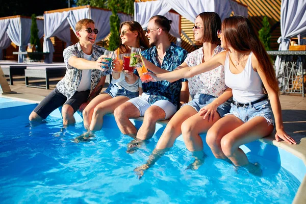 Grupo de amigos se divertindo na festa na piscina copos clinking com coquetéis frescos sentados à beira da piscina no dia ensolarado de verão. As pessoas brindam a beber bebidas na villa de luxo em férias tropicais. — Fotografia de Stock