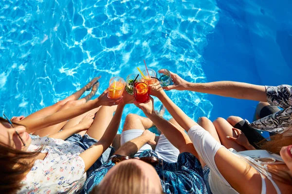 Vista superior dos amigos copos clinking com coquetéis coloridos frescos sentados à beira da piscina no dia ensolarado de verão. As pessoas brindam a beber bebidas na luxuosa festa à beira da piscina em férias tropicais. — Fotografia de Stock