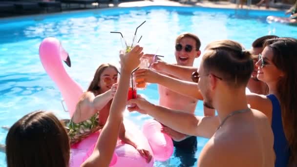 Multiracial group of friends toasting, clinking glasses with cocktails at swimming pool party. Happy young people in swimwear dancing, clubbing with inflatable flamingo, mattresses in luxury resort. — Stock Video