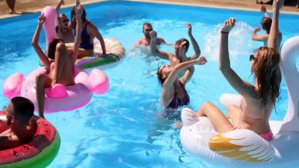 Friends have party in a private villa swimming pool. Happy young people in swimwear splashing water, dancing with floaties and inflatable mattress in luxury resort on sunny day. Slow motion. — Stock Video