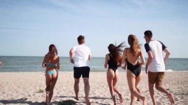 Bir grup mutlu arkadaş deniz suyuna koşuyor ve kumsalda zıplıyorlar. Neşeli insanlar yaz güneşli bir günde deniz kenarında eğleniyorlar. Genç erkekler ve kadınlar okyanusa yüzmeye giderler. 120 fps yavaş çekim.