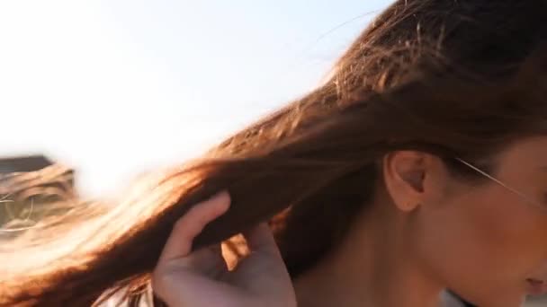 Schließen Sie die Rückansicht der braunen Frauenhaare zarte Bewegung in der Luft in Zeitlupe 120 fps. Brünette Frau mit Sonnenbrille am Sandstrand. Der Wind bläst luftige Haare, die bei Sonnenuntergang in der Sonne glänzen. Makrovideo. — Stockvideo