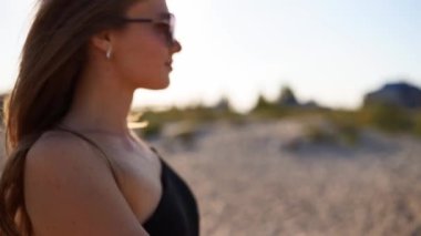 Güneşli bir tropikal plajda yürüyen mayo ve güneş gözlüklü çekici sarışın kadın. Uzun saçlı, bikinili güzel kız kumlu deniz kıyısında ağır çekimde yürüyüşün tadını çıkarıyor. Güneş patlamaları.