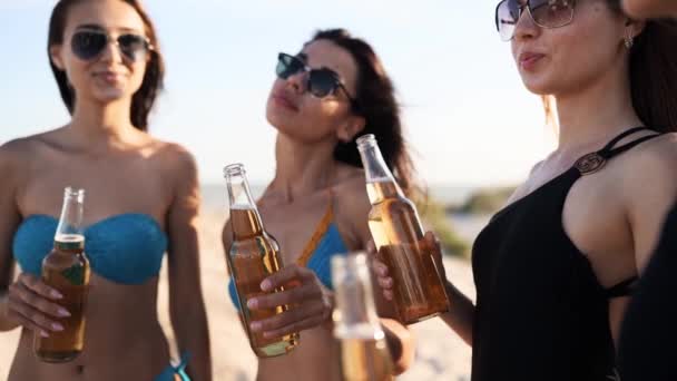 Bonitas mujeres en bikini divirtiéndose brindando por la bebida y relajándose en la playa de arena al atardecer. Chicas equipadas en trajes de baño, gafas de sol colgando en la orilla del mar con botellas de cerveza. Las hembras jóvenes beben limonada. — Vídeos de Stock