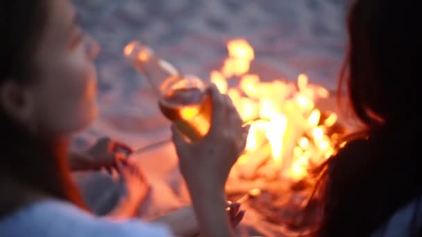 Junge Frau trinkt Bier aus Glasflasche und sitzt mit Freunden am Lagerfeuer und frittiert Würstchen am Sandstrand. Mädchen mit Getränken entspannen am Lagerfeuer in der Abenddämmerung. Weibliches Chillen mit Limonade. — Stockvideo