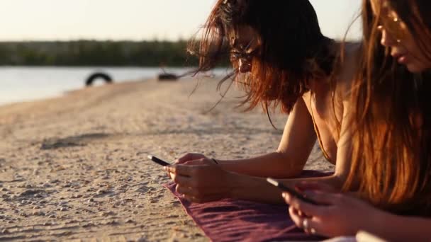 Grupa milenijnych kobiet korzystających ze smartfonów leżących razem na ręczniku plażowym w pobliżu morza w letni zachód słońca. Młodzi ludzie uzależnieni od telefonów komórkowych. Zawsze połączone pokolenie komunikuje się przez internet. — Wideo stockowe