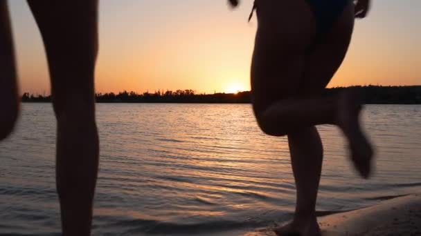 Chicas mojadas felices en bikini corren al mar, juegan salpicaduras de agua entre sí en la puesta del sol. Las amigas se divierten haciendo salpicaduras en el lago. Las siluetas de las mujeres jóvenes van a nadar, bañarse en el estanque en el atardecer. Lento. — Vídeo de stock