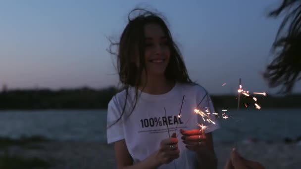 Amigas caminando, bailando, divirtiéndose en la fiesta nocturna en la playa con bengalas en las manos. Jóvenes adolescentes festejando en la playa con fuegos artificiales, luces de bengala. Las niñas en cámara lenta, steadycam disparo. — Vídeos de Stock