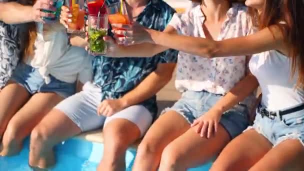 Przyjaciele bawią się na imprezie przy basenie brzęcząc szklankami z kolorowymi koktajlami siedzącymi przy basenie w słoneczny letni dzień. Ludzie piją napoje ze stopami w wodzie w willi wakacyjnej. — Wideo stockowe