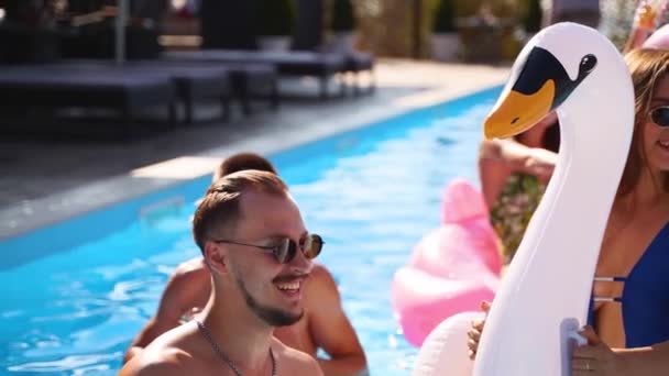 Freunde feiern mit Cocktails im Pool der Ferienvilla. Fröhliche junge Leute in Badebekleidung tanzen, Clubbing mit aufblasbarem Flamingo, Schwan, Matratze im Luxus-Resort an sonnigen Tagen. Zeitlupe — Stockvideo