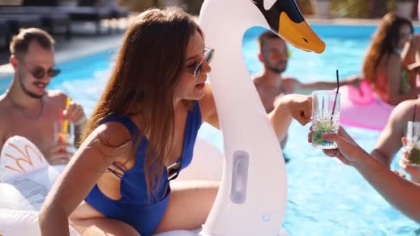Freunde feiern mit Cocktails im Pool der Ferienvilla. Fröhliche junge Leute in Badebekleidung tanzen, Clubbing mit aufblasbarem Flamingo, Schwan, Matratze im Luxus-Resort an sonnigen Tagen. Zeitlupe — Stockvideo