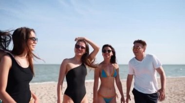 Bir grup arkadaş güneşli sahilde yürüyor. Genç bronzlaşmış neşeli melez insanlar yazın deniz kenarında yürüyor, sohbet ediyor ve gülüyorlar. Erkekler ve kadınlar mayo ve bikini giyiyor. Yaşam tarzı ağır çekim.