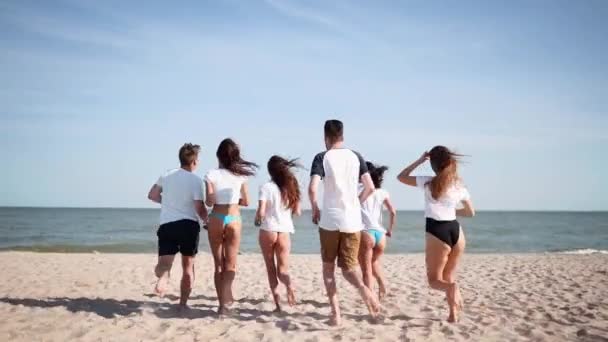 Groep gelukkige vrienden kleden zich uit in het zeewater en gooien shirts op het strand. Vrolijke mensen die zich vermaken aan de kust bij de oceaan. Jonge mannen en vrouwen gaan zwemmen en baden. 120 fps slow motion. — Stockvideo