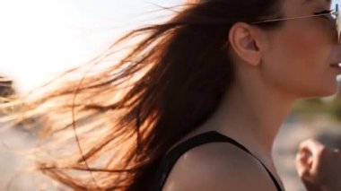 Kahverengi kadın saçının yavaşça hareket edişini yakından izleyin. 120 fp. Kumsalda yürüyen güneş gözlüklü esmer kadın. Rüzgâr, günbatımında güneşte parlayan havadar saçlar savurur. Makro video.