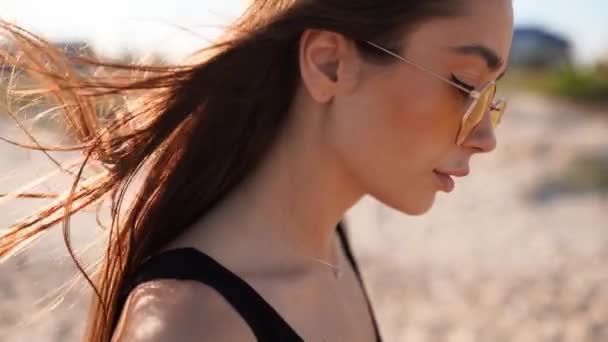 Vista posteriore ravvicinata del movimento tenero dei capelli della donna marrone nell'aria al rallentatore 120 fps. Bruna donna in occhiali da sole a piedi sulla spiaggia di sabbia. Il vento soffia capelli ariosi che splendono al sole al tramonto. Macro video. — Video Stock