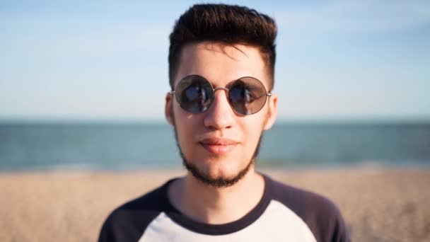 Portret van een jonge man die kijkt en lacht naar een camera die op het strand staat met een zee op de achtergrond. gelukkig guy in zonnebril kijken naar camera ontspannen op oceaan kust op zonnige dag buiten. — Stockvideo