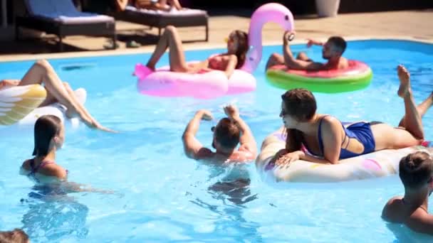 Os amigos que refrigeram na piscina privada da casa de campo, encontram-se no sol no flamingo inflável, cisne, flutuadores. Os jovens relaxam na festa no resort de luxo no dia ensolarado. Meninas de biquíni tomando banho de sol. Movimento lento. — Vídeo de Stock