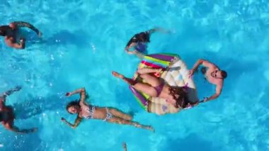 Havuzda, şişme flamingo, kuğu ve şiltelerle parti veren arkadaşlar. Mutlu gençler güneşli bir günde lüks bir tatil köyünde dinleniyorlar. Yukarıdan bak. Bikinili kızlar güneşte güneşleniyor..
