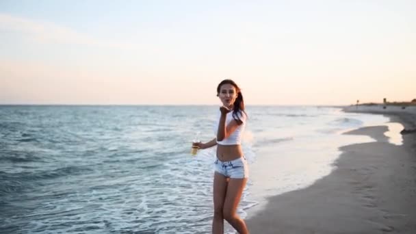 Vacker kvinna som går längs havet på en sandstrand och har roligt med ölflaskan. Attraktivt utrustad flicka skickar luft kyss, njuter av sommarsemester nära havet vågor med dryck. Följ mig. Långsamma rörelser. — Stockvideo
