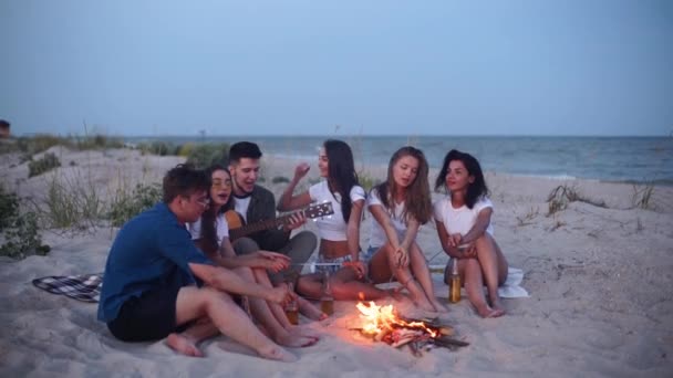 Przyjaciele siedzą przy ognisku, piją piwo, śpiewają na gitarze, smażą kiełbaski na piaszczystej plaży. Młody mieszany rasa grupa mężczyzn i kobiet z napojów śpiew wzdłuż, brodaty facet gra na gitarze na imprezie w zmierzchu. — Wideo stockowe