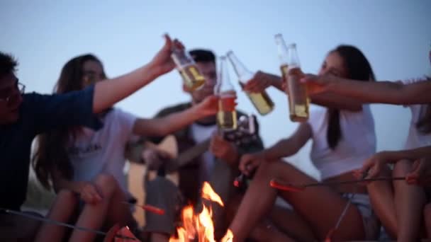 Φίλοι κάθονται γύρω από τη φωτιά ζητωκραυγάζοντας και ψήνοντας μπουκάλια μπύρας, παίζοντας κιθάρα, και τηγανίζοντας λουκάνικα στην αμμώδη παραλία. Νεαρή ομάδα ανδρών και γυναικών πίνουν ποτά, τραγουδώντας. Ο τύπος παίζει κιθάρα το σούρουπο.. — Αρχείο Βίντεο