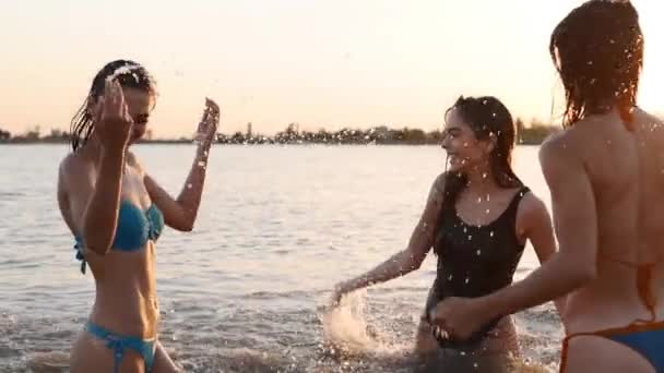 Ευτυχισμένα βρεγμένα κορίτσια με μπικίνι τρέχουν στη θάλασσα και παίζουν νερό ο ένας στον άλλο το ηλιοβασίλεμα. Χαρούμενες φίλες διασκεδάζουν κάνοντας πιτσίλισμα στη λίμνη. Νεαρές γυναίκες πάνε για κολύμπι και μπάνιο στη λίμνη. Αργή κίνηση. — Αρχείο Βίντεο