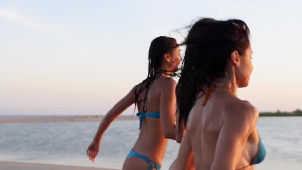Glada våta tjejer i bikini som springer ut i havsstänk vid solnedgången. Glada kvinnliga vänner som har kul på stranden nära dammen. Unga kvinnor badar och badar i sjön. Sprickor i långsamma rörelser. — Stockvideo
