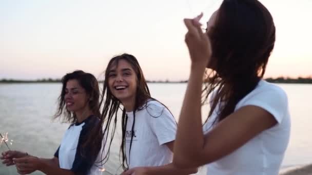 Des amies marchant, dansant, s'amusant à la soirée au bord de la mer avec des étincelles dans les mains. Jeunes adolescentes faisant la fête sur la plage avec feu d'artifice, lumières du bengale. Les filles au ralenti, steadycam shot. — Video