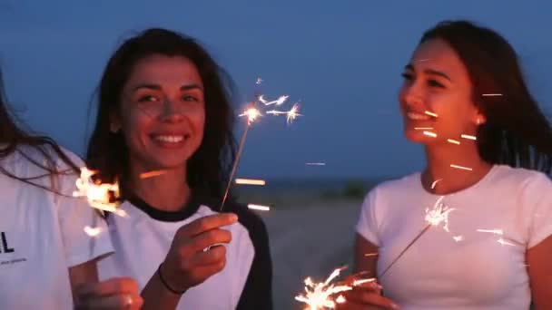 Amigas caminando, bailando, divirtiéndose en la fiesta nocturna en la playa con bengalas en las manos. Jóvenes adolescentes festejando en la playa con fuegos artificiales, luces de bengala. Las niñas en cámara lenta, steadycam disparo. — Vídeo de stock