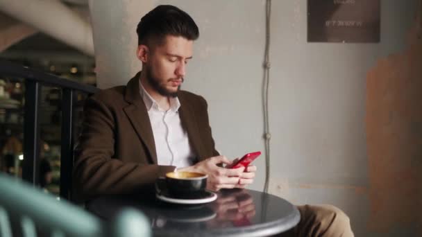 カフェに座っているスマートフォンを使用してクラスの若い男。コーヒーショップでソーシャルネットワークアプリを閲覧する男。オフィスワーカーはコーヒーを飲みながら休憩している。屋内シーン. — ストック動画