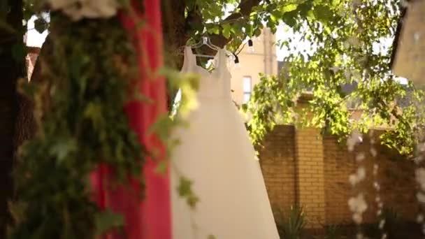Svatební šaty visí na stromě na rustikální svatbě. Venkovský dekor s růžovými kyticemi, girlandami a látkou. — Stock video