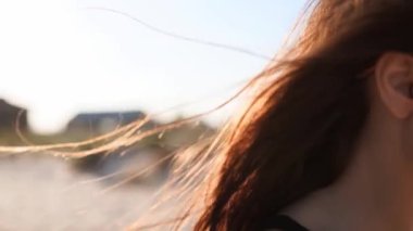 Kahverengi kadın saçının yavaşça hareket edişini yakından izleyin. 120 fp. Kumsalda yürüyen güneş gözlüklü esmer kadın. Rüzgâr, günbatımında güneşte parlayan havadar saçlar savurur. Makro video.
