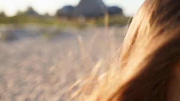 Vista da vicino di donna bionda movimento tenero dei capelli in aria al rallentatore 120 fps. Donna marrone chiaro in occhiali da sole che cammina sulla spiaggia. Il vento soffia capelli ariosi che splendono al sole al tramonto. Macro video. — Video Stock