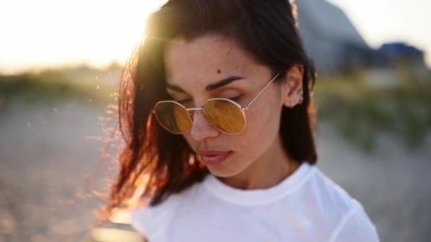 Ritratto di giovane donna che guarda la macchina fotografica in piedi sulla spiaggia con un mare sullo sfondo. Attraente ragazza in t-shirt bianca e occhiali da sole alza gli occhi. Il vento soffia ariosi capelli castani che brillano nei razzi del sole. — Video Stock