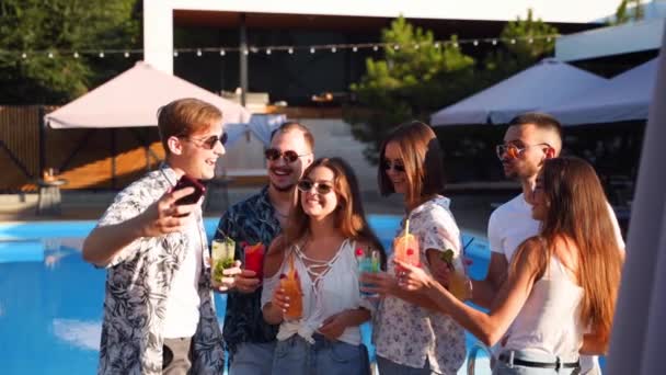 Amici sorridenti scattare selfie con smartphone alla festa in piscina con cocktail colorati in piedi a bordo piscina nella soleggiata giornata estiva. Uomo che scatta foto in villa di lusso in vacanza tropicale. Rallentatore. — Video Stock