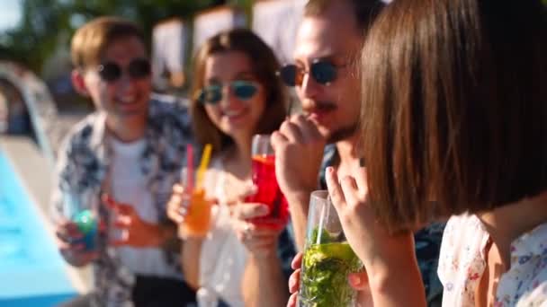 Grupo de amigos divirtiéndose en la fiesta junto a la piscina tintineando vasos con cócteles frescos sentados junto a la piscina en el soleado día de verano. La gente brinda bebiendo bebidas en una villa de lujo en vacaciones tropicales. — Vídeo de stock