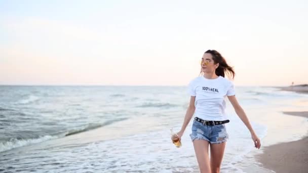 Pretty woman walking along seaside on sandy beach having fun with beer bottle. Attractive fitted girl raises hands, enjoys fredom and summer vacation near ocean shore, drinking beverage. Slow motion. — Stock Video