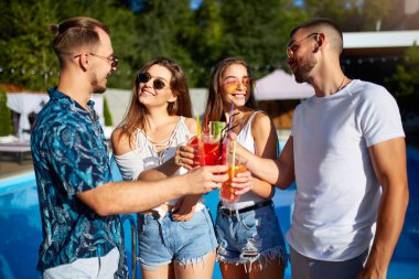 Bir grup arkadaş, havuz başında yaz partisinde bardakları tokuşturup güneşli bir günde havuz kenarında kokteyl içiyorlar. İnsanlar tropikal tatillerde lüks villada taze meyve suyu içerek kadeh kaldırıyorlar..