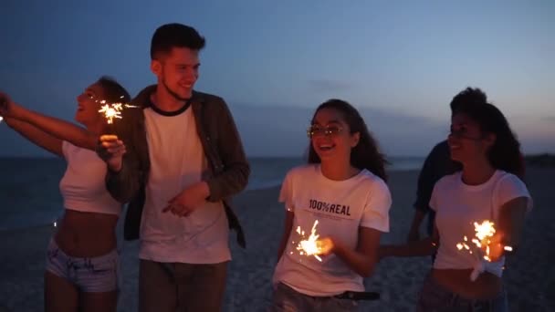 Amici che camminano, ballano, si divertono alla festa notturna al mare con scintille in mano. Giovani adolescenti che festeggiano sulla spiaggia con fuochi d'artificio e luci del bengala. Scatto steadycam rallentato. — Video Stock