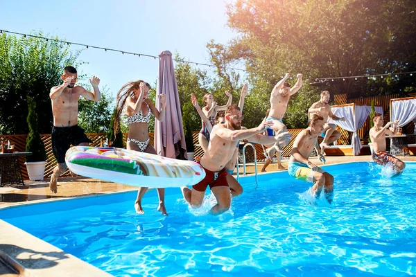 Щасливі друзі стрибають і бризкають в басейні з надувними поплавками в розкішному курорті. Молоді люди в купальнику проводять вечірку на приватній віллі в сонячний день. Гарячі дівчата стрибають у воду . — стокове фото