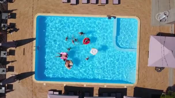 Luchtfoto van vrienden die feesten in het zwembad met opblaasbare flamingo, zwaan, matras. Gelukkige jongeren zwaaien handen naar drone camera op luxe resort. Zicht van bovenaf. Meisjes in bikini zonnebaden. — Stockvideo