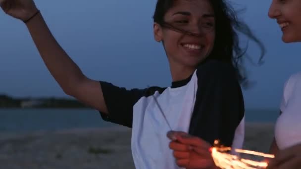 Девушки ходят, танцуют, веселятся на ночной вечеринке на берегу моря с бенгальскими огнями в руках. Молодые девушки-подростки тусуются на пляже с фейерверком, бенгальскими огнями. Девушки в замедленной съемке, устойчивый кадр. — стоковое видео