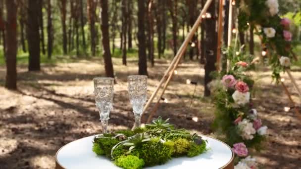 お客様のための花やキャンドルや白い椅子で飾られたbohemian tipiアーチの横にある白いテーブルの上に草や苔のワイングラス、結婚指輪の寝具. — ストック動画
