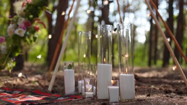 Candles burning in glass vases, flasks on carpet near bohemian tipi arch decorated with roses and flowers wrapped in fairy lights on outdoor wedding ceremony venue in pine forest. Boho rustic decor. — Stock Video