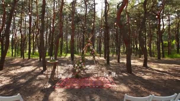 Belle décoration bohème en arc de tipi sur le lieu de cérémonie de mariage en plein air dans la forêt de pins avec des cônes. Chaises, compositions floristiques de fleurs de roses, tapis, lumières de fées à cordes. Mariage rural d'été. — Video