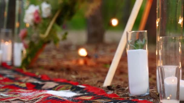 Candles burning in glass vases, flasks on carpet near bohemian tipi arch decorated with roses and flowers wrapped in fairy lights on outdoor wedding ceremony venue in pine forest. Boho rustic decor. — Stock Video
