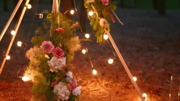Arco de madera de tipi bohemio decorado con velas encendidas, rosas y hierba de pasto, envuelto en luces de hadas iluminación en el lugar de la ceremonia de boda al aire libre en el bosque de pinos por la noche. Bulbos guirnalda brilla. — Vídeo de stock