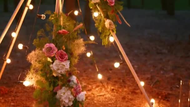 Arco de madeira boêmio tipi decorado com velas em chamas, rosas e grama pampass, envolto em luzes de fadas iluminação no local da cerimônia de casamento ao ar livre em pinhal à noite. Lâmpadas guirlanda brilha. — Vídeo de Stock