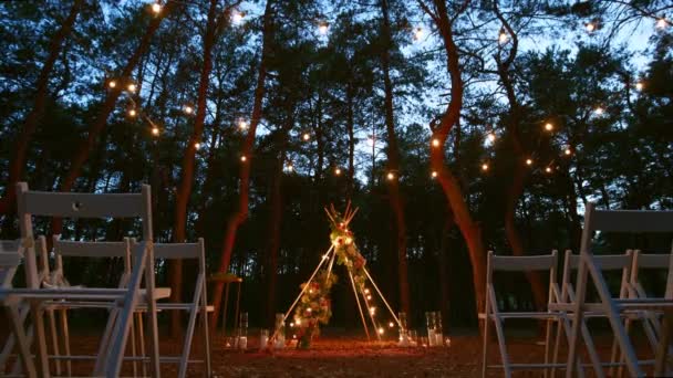 Festive string lights illumination on boho tipi arch decor on outdoor wedding ceremony venue in pine forest at night. Vintage string lights bulb garlands shining above chairs at summer rural wedding. — Stock Video